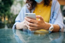 Eine Frau mit Mundschutz hält ein Mobiltelefon in den Händen